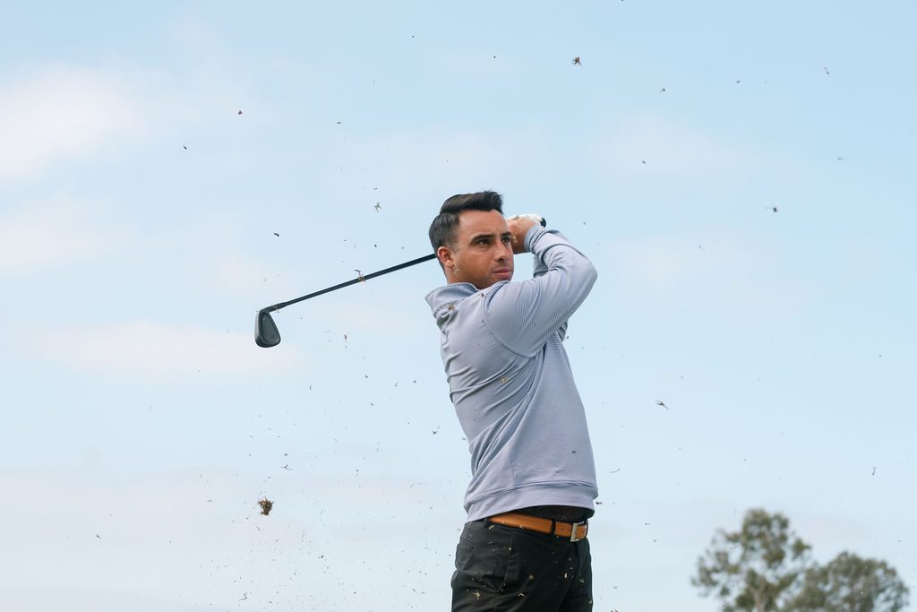 A golfer holds his finish as turf flies against a blue sky