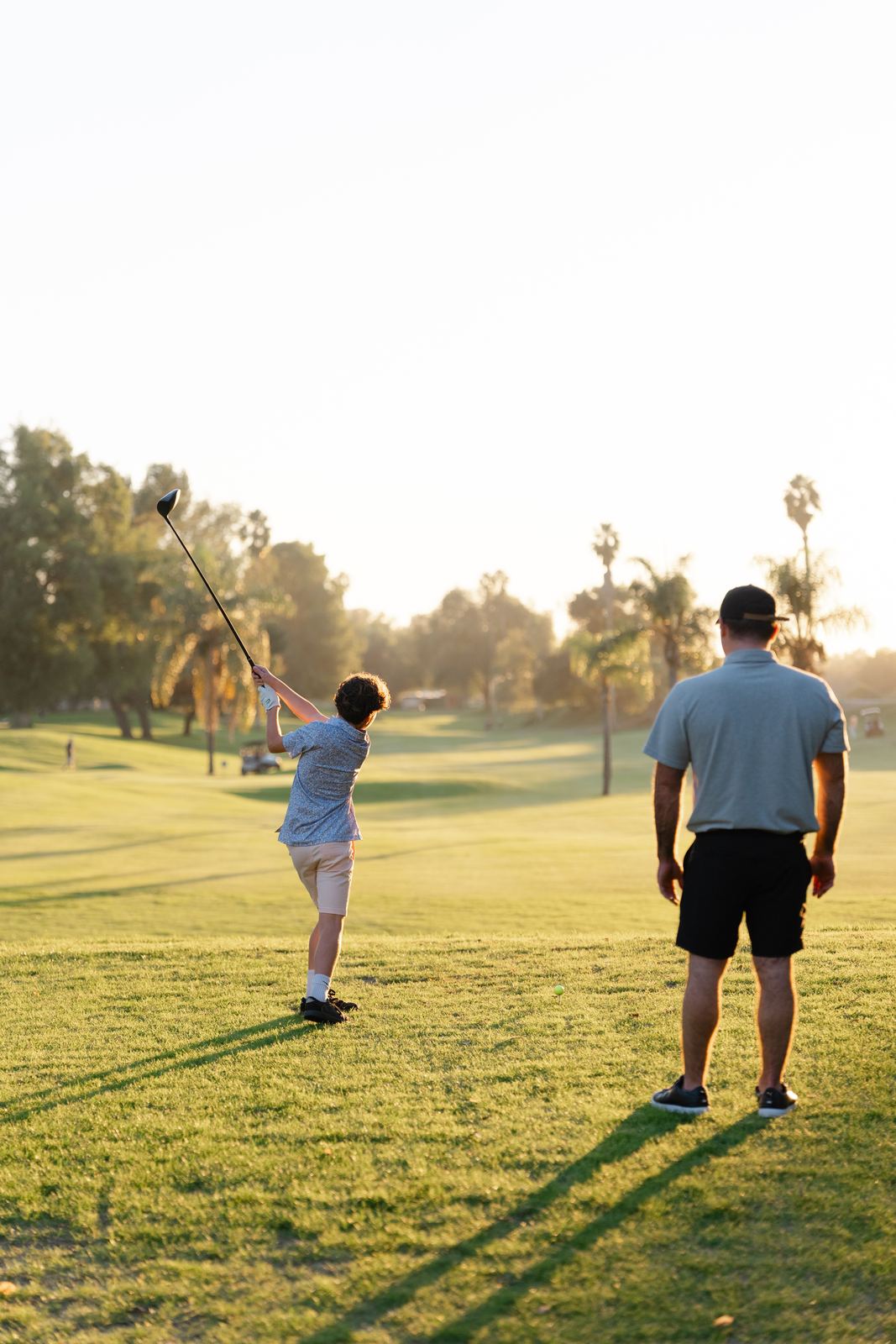 A kid tees off while his dad watches at golden hour